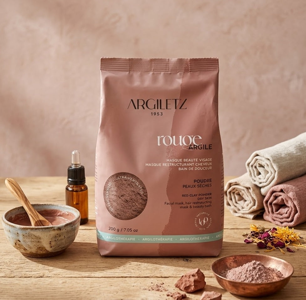 Un sachet d'Argiletz 1953 Argile Rouge Ultra-Ventilée Poudre Peaux Sèches de 200g est disposé sur une table en bois rustique. Le sachet, au centre, est entouré d'accessoires de spa, notamment un bol en céramique avec un masque d'argile préparé et une cuillère en bois, un bol en cuivre contenant de la poudre d'argile brute, des flacons d'huile, des serviettes en lin roulées et des fleurs séchées, créant une scène de rituel de beauté bien-être pour le visage et les cheveux.
