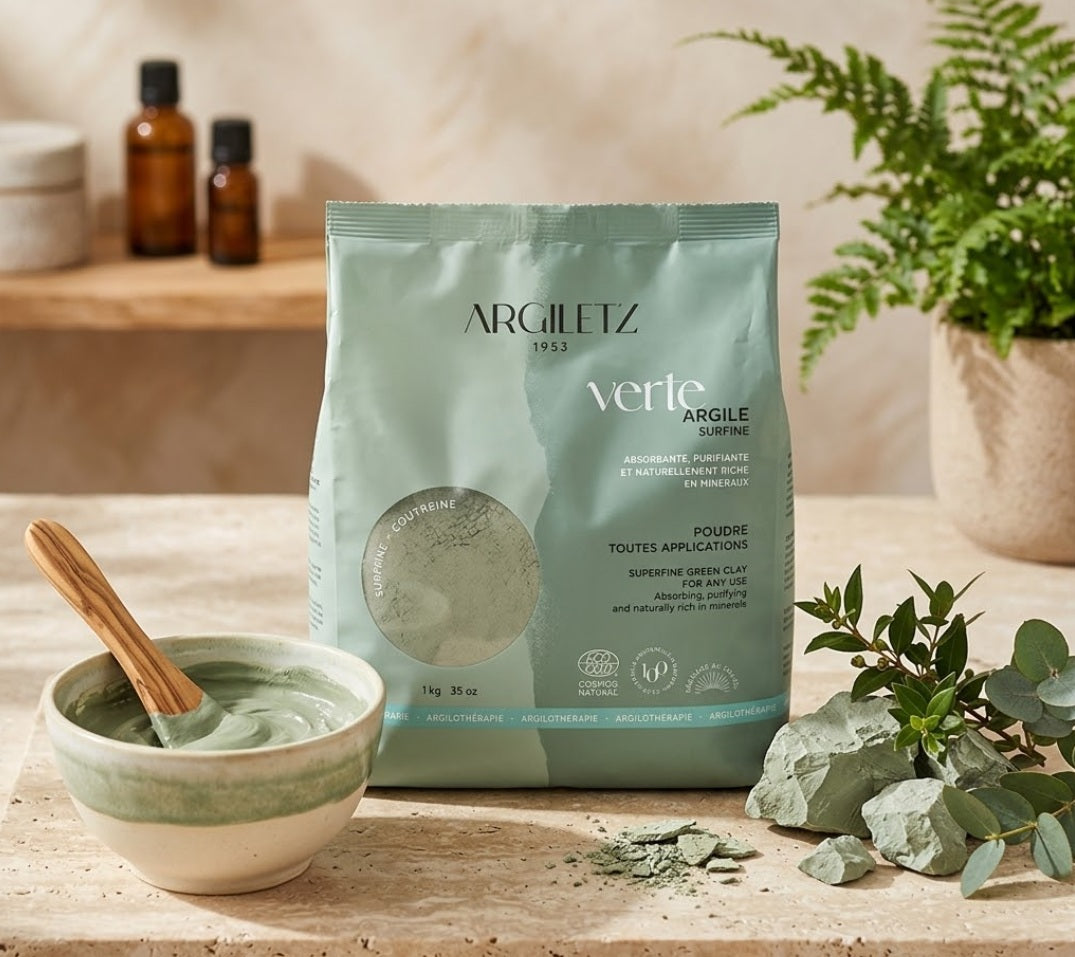 Mise en scène de soin naturel avec un sachet d'Argile Verte Surfine Argiletz 1kg au centre. À gauche, un bol contient de la poudre d'argile verte préparée avec une spatule en bois. À droite, des morceaux d'argile brute sont disposés avec des branches d'eucalyptus. L'arrière-plan montre une étagère en bois avec des flacons de soin flous et une plante verte en pot, dans une ambiance de salle de bain de spa. Ce produit d'argilothérapie est 100% naturel et certifié ECOCERT.