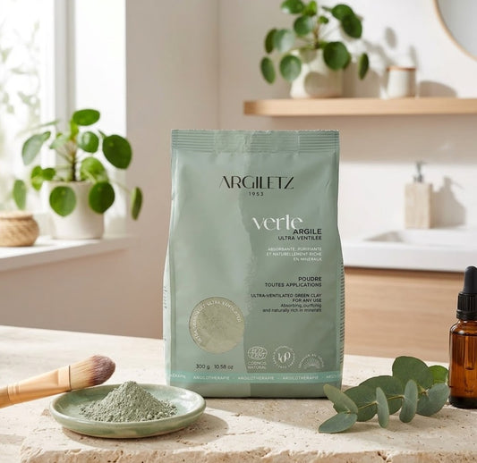 Un sachet de sachet de "verte Argile ULTRA VENTILÉE" d'ARGILÉTZ 1953, de couleur gris-vert, posé sur une surface en pierre texturée dans une salle de bain lumineuse et épurée. À côté, une petite coupelle contient de la poudre d'argile verte, un pinceau de maquillage à manche en bois, une branche d'eucalyptus fraîche et un flacon de sérum en verre ambré. En arrière-plan, des plantes vertes en pot et des étagères en bois créent une ambiance de bien-être naturel.