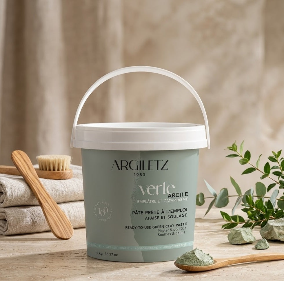Photographie de produit attrayante d'un seau de pâte d'argile verte prête à l'emploi Argiletz, 1kg, posé sur un comptoir de travertin dans une salle de bain de style spa. Le seau est entouré de serviettes en lin texturé, d'une spatule en bois d'olivier, et de branches fraîches d'eucalyptus et de myrte. Tous les textes et logos de l'emballage sont nets et lisibles.