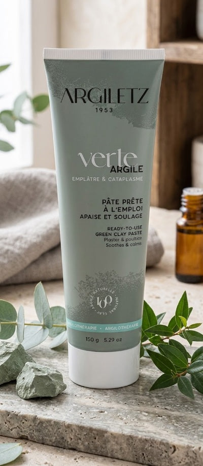 Photographie de mise en scène produit d'un tube de 150g de pâte d'argile verte Argiletz 1953 'Verte Argile'. L'emballage vert bleuté mat est décoré de motifs d'eucalyptus et porte les mentions 'Pâte prête à l'emploi, Apaise et soulage'. Le tube est posé sur un comptoir en marbre gris, entouré de branches d'eucalyptus fraîches, de morceaux d'argile brute, d'une serviette en lin et d'un flacon d'huile essentielle ambré en arrière-plan.